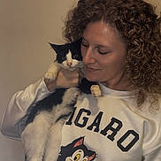 Figaro is registered to the contest to win money with this photo: woman, cat, curly_hair, white_sweatshirt, cartoon_cat, indoor, pet, black_and_white_cat, smile, affection, holding, portrait, cute, feline, human, casual_clothing, young_adult, animal, friendly, love