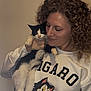 woman, cat, curly_hair, white_sweatshirt, cartoon_cat, indoor, pet, black_and_white_cat, smile, affection, holding, portrait, cute, feline, human, casual_clothing, young_adult, animal, friendly, love