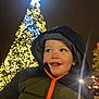 child, toddler, smile, hood, jacket, zipper, night, christmas_tree, lights, holiday, festive, outdoor, portrait, face, winter_clothing, warm, happy, glow, bokeh, celebration