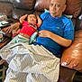 baby, man, sleeping, couch, leather, blanket, living_room, relaxing, child, adult, casual_clothing, red_shirt, blue_shirt, home, resting, comfort, pillow, hand, indoor, nap