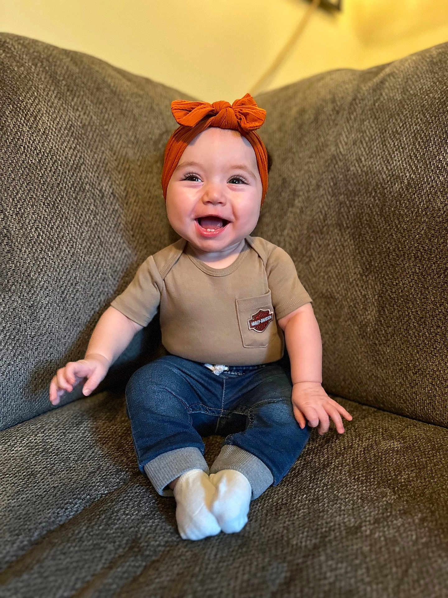 Eleanor is registered to the contest to win money with this photo: baby, infant, smiling, headband, bow, couch, sofa, sitting, portrait, happy, onesie, jeans, socks, living_room, cute, chubby_cheeks, hands, face, cozy, furniture