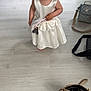 bag, barefoot, child, curious, dress, floor, furniture, girl, home, indoor, light, pacifier, person, pigtails, shelf, standing, toddler, toy, white_dress, wooden_floor