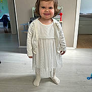 Catalina a rejoint le concours — aidez-le/la à gagner de superbes lots ! cardigan, child, cute, furniture, girl, hair_bow, home, indoor, light_floor, person, playful, portrait, room, smile, standing, toddler, tongue_out, toy, white_dress, young_child