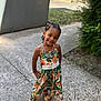 child, girl, smile, braided_hair, dress, sandals, outdoor, sidewalk, greenery, confident_pose, flower_pattern, happy, cute, portrait, sunlight, nature, fashion, accessories, young, casual