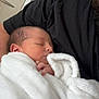 newborn, baby, blanket, sleeping, person, black_shirt, indoor, closeup, soft_texture, peaceful, infant, resting, wrapped, portrait, human, skin, hand, head, face, cozy