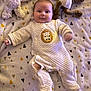 baby, infant, onesie, blanket, stuffed_animal, plush_toy, smiling, face, person, soft_texture, quilted, lying_down, cute, indoors, child, animal_toy, sleepwear, cozy, patterned_blanket, warm_lighting