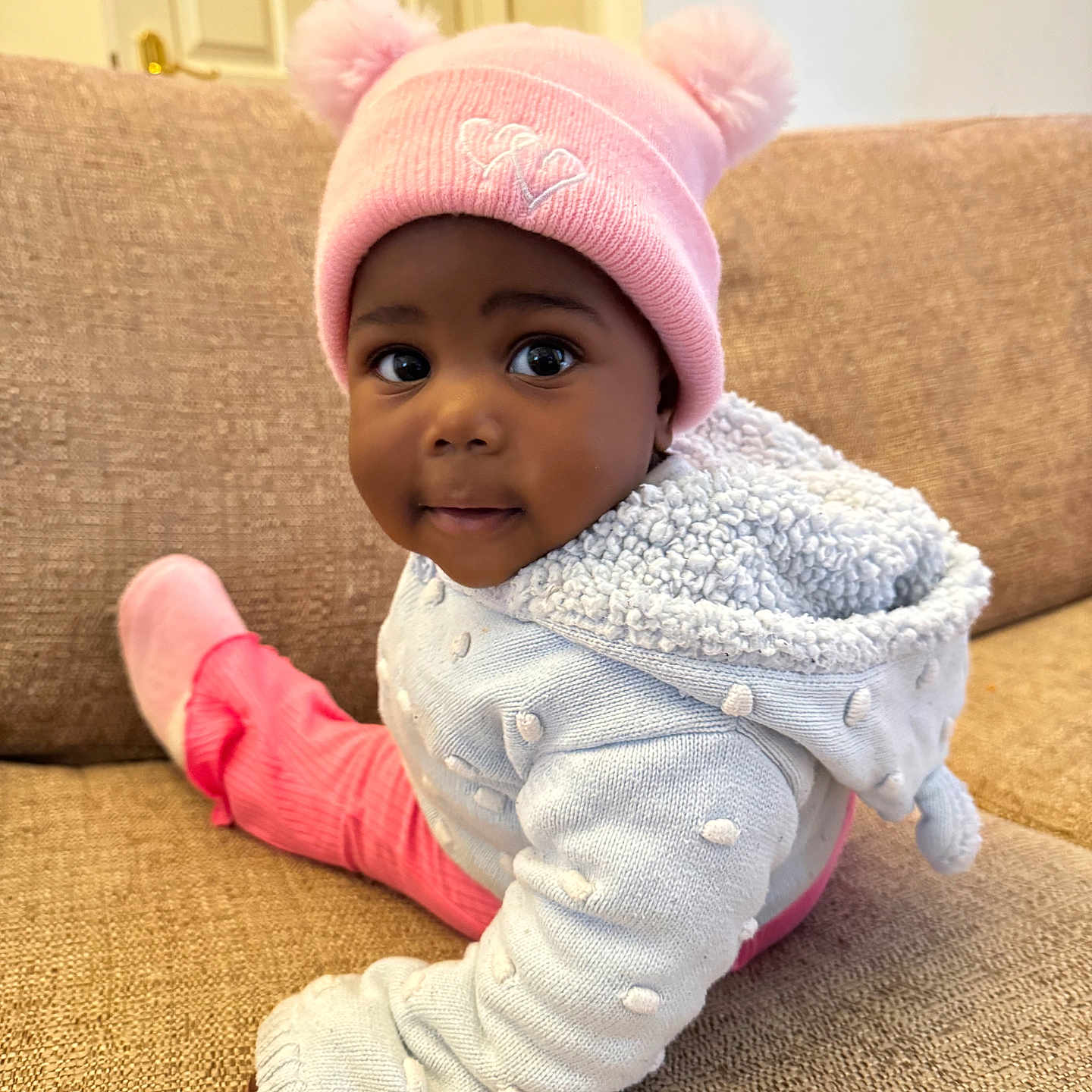 Chantel is registered to the contest to win money with this photo: baby, beige_couch, child, close_up, couch, cozy, cute, face, hat, indoor, infant, looking, person, pink_hat, pom_poms, portrait, sitting, sweater, warm_clothing, white_sweater