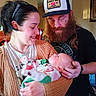 woman, man, baby, indoor, family, holding, smiling, beard, hat, casual_clothing, love, care, cozy, portrait, newborn, affection, warmth, head_support, sweater, peaceful
