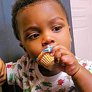O'Mari is registered to the contest to win money with this photo: toddler, child, cupcake, food, indoor, face, hand, clothing, pattern, frosting, cabinet, curly_hair, snack, eating, young_child, portrait, cute, expression, colorful, standing