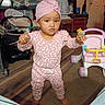 toddler, child, baby, standing, portrait, pink_outfit, head_wrap, serious_expression, raised_hands, indoor, living_room, toy_walker, baby_gear, stroller, crib, wood_floor, carpet, furniture, playtime, photography