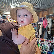 Lincoln joined the competition — help win amazing prizes! baby, child, straw_hat, yellow_onesie, drooling, tattoo, adult_hand, indoor, cluttered_background, fan, shelf, clothing, person, face, holding, curious, blue_eyes, furniture, room, portrait