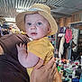 baby, child, straw_hat, yellow_onesie, drooling, tattoo, adult_hand, indoor, cluttered_background, fan, shelf, clothing, person, face, holding, curious, blue_eyes, furniture, room, portrait