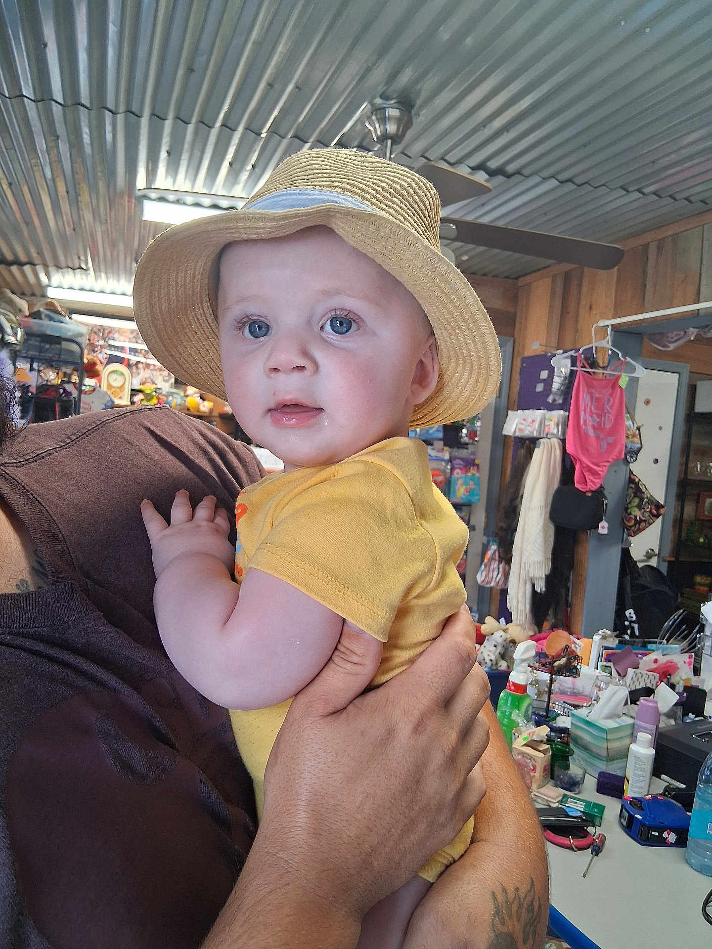 Lincoln joined the competition — help win amazing prizes! baby, child, straw_hat, yellow_onesie, drooling, tattoo, adult_hand, indoor, cluttered_background, fan, shelf, clothing, person, face, holding, curious, blue_eyes, furniture, room, portrait