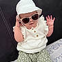 accessory, baby, bed, bedding, casual, child, cute, fashion, green_pants, hand, hat, headwear, indoor, infant, patterned_pants, portrait, sitting, smiling, sunglasses, white_clothing