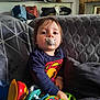 baby, child, colorful, couch, cute, furniture, gray_blanket, home, indoor, living_room, pacifier, person, playtime, portrait, relaxed, seated, superman_shirt, toddler, toys, young_child