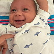 Emiliano is registered to the contest to win money with this photo: baby, infant, smile, face, hand, clothing, onesie, shark_pattern, happy, cute, human, person, skin, fingers, lying_down, bed, textile, closeup, expression, joy