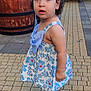 child, toddler, dress, blue, floral_pattern, hair_clips, sandals, outdoor, paved_walkway, wooden_barrel, bench, curious, standing, person, young_child, portrait, daylight, side_view, expression, casual_wear