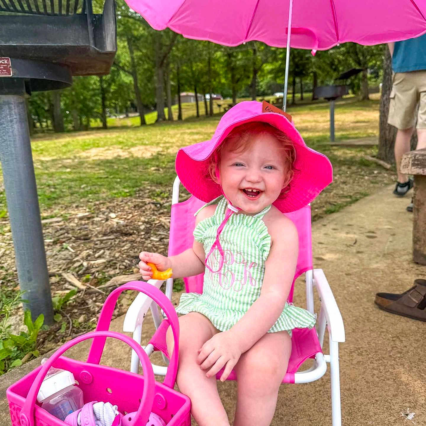 Wrenliegh Kate is registered to the contest to win money with this photo: baby_bottle, child, footwear, grass, green_outfit, happy, outdoor, park, person, picnic_area, pink_chair, pink_hat, pink_umbrella, seat, smiling, snack, sunny, toddler, toy_bag, trees
