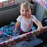 Kylee is registered to the contest to win money with this photo: toddler, child, playpen, smile, happy, indoors, blanket, toy, hair, pigtails, clothing, person, standing, cute, colorful, fabric, window, daylight, expression, home