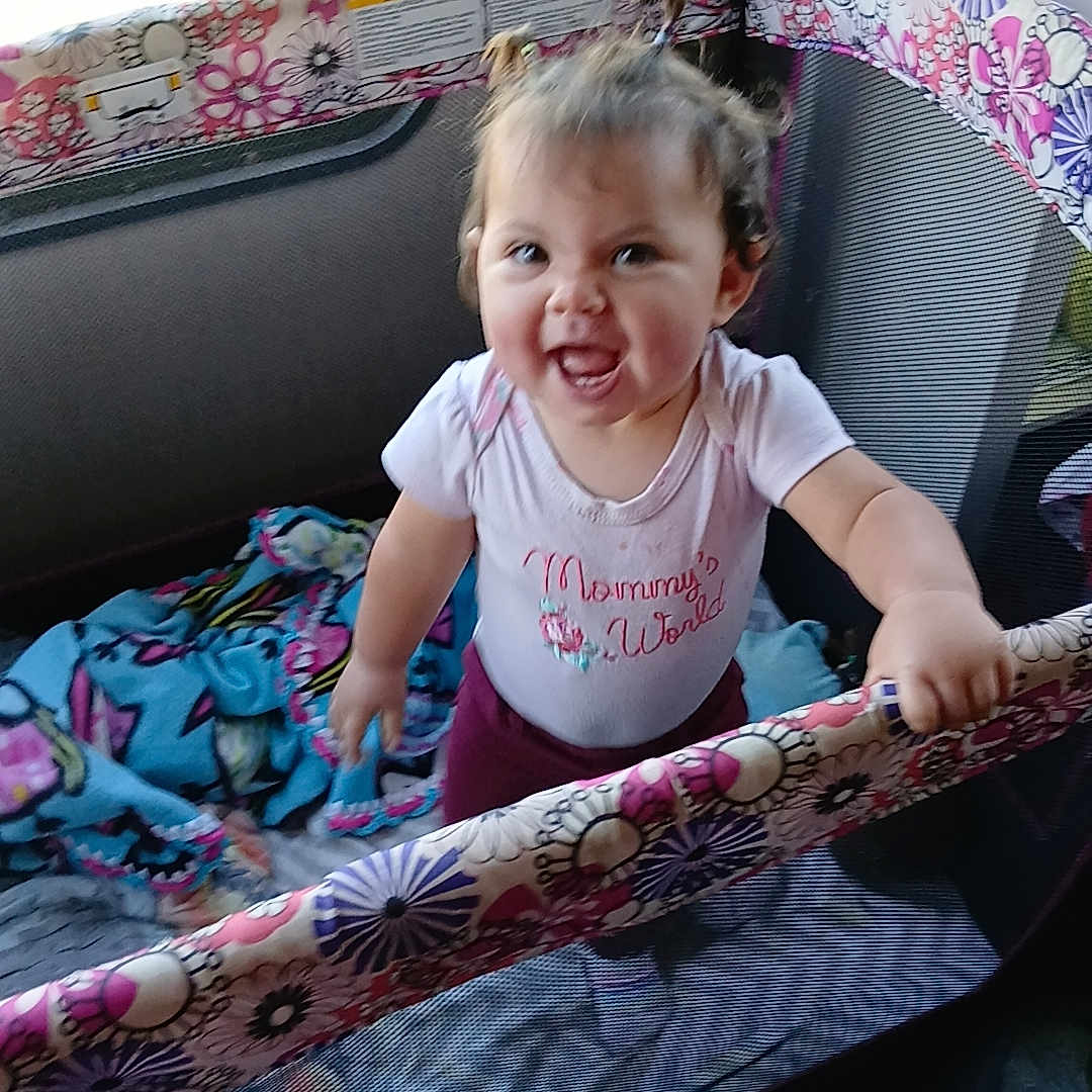 Kylee is registered to the contest to win money with this photo: toddler, child, playpen, smile, happy, indoors, blanket, toy, hair, pigtails, clothing, person, standing, cute, colorful, fabric, window, daylight, expression, home