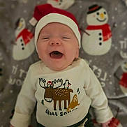 Lincoln is registered to the contest to win money with this photo: baby, smiling, santa_hat, sweater, moose, penguin, blanket, snowman, holiday, christmas, cute, infant, happy, clothing, face, child, lying_down, indoor, festive, cozy