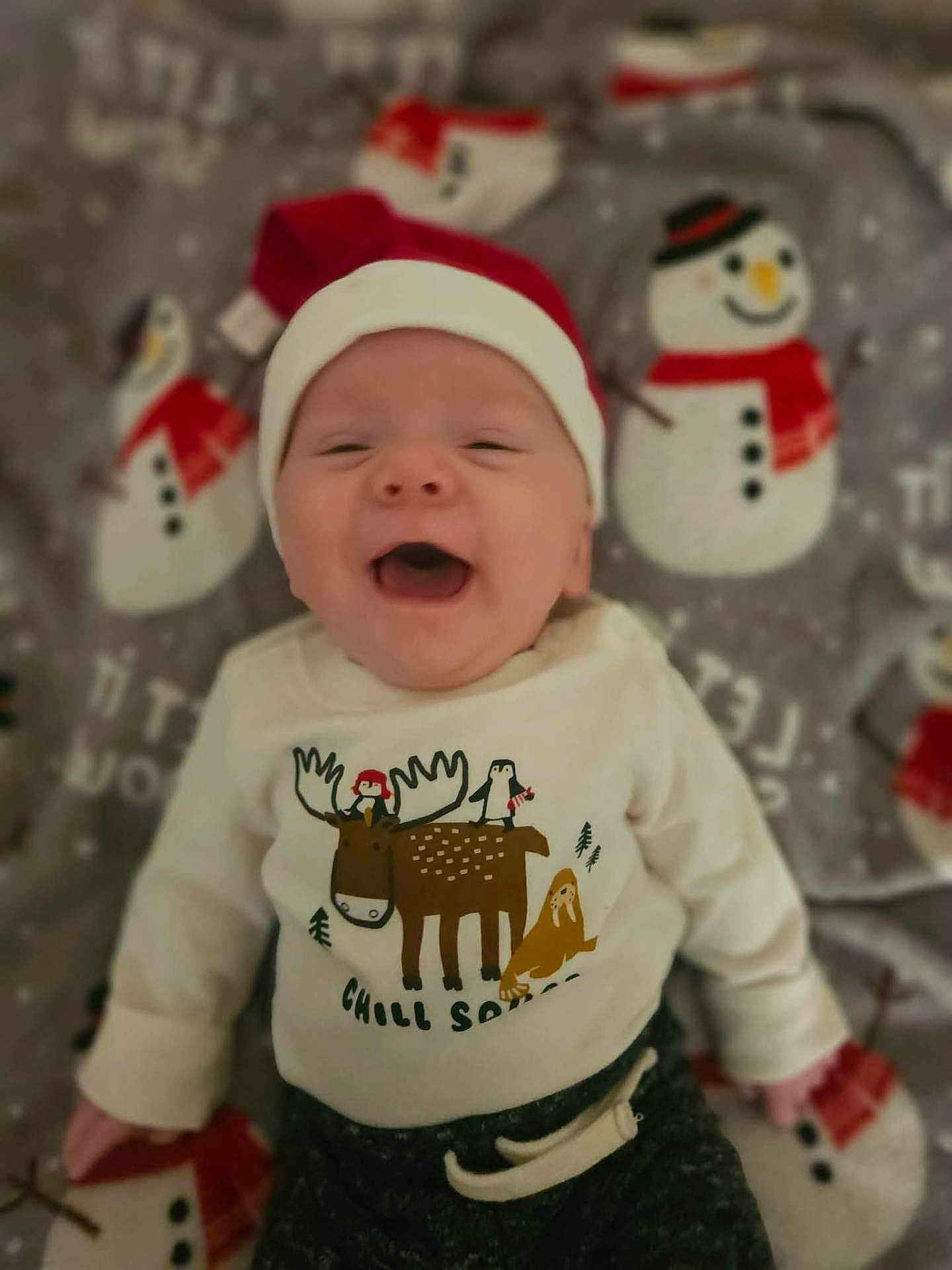 Lincoln is registered to the contest to win money with this photo: baby, smiling, santa_hat, sweater, moose, penguin, blanket, snowman, holiday, christmas, cute, infant, happy, clothing, face, child, lying_down, indoor, festive, cozy