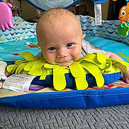 Gunter is registered to the contest to win money with this photo: baby, blanket, blue_cushion, book_shelf, child, colorful, face, floor, furniture, happy, head, indoor, person, play_mat, portrait, smiling, soft_toys, toy_fish, toy_octopus, yellow_cushion