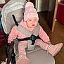 baby, child, stroller, pink_clothing, hat, pom_pom, boots, indoor, wooden_floor, door, straps, seat, curious_expression, face, person, baby_toy, warm_clothing, hoodie, cushion, baby_seat