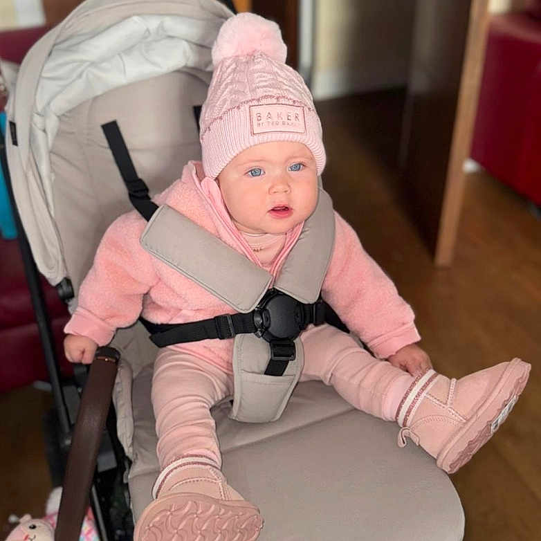 Sharon-Antonia joined the competition — help win amazing prizes! baby, baby_seat, baby_toy, boots, child, curious_expression, cushion, door, face, hat, hoodie, indoor, person, pink_clothing, pom_pom, seat, straps, stroller, warm_clothing, wooden_floor
