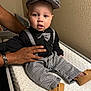 baby, child, person, infant, hat, bow_tie, suspenders, black_shirt, houndstooth_pattern, pants, moccasins, hand, changing_pad, indoor, wall, curious_expression, sitting, support, fashion, portrait