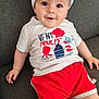 apparel, baby, casual, child, clothing, couch, cute, face, happy, hat, headwear, indoor, infant, person, playful, red_shorts, shorts, sitting, smiling, tshirt