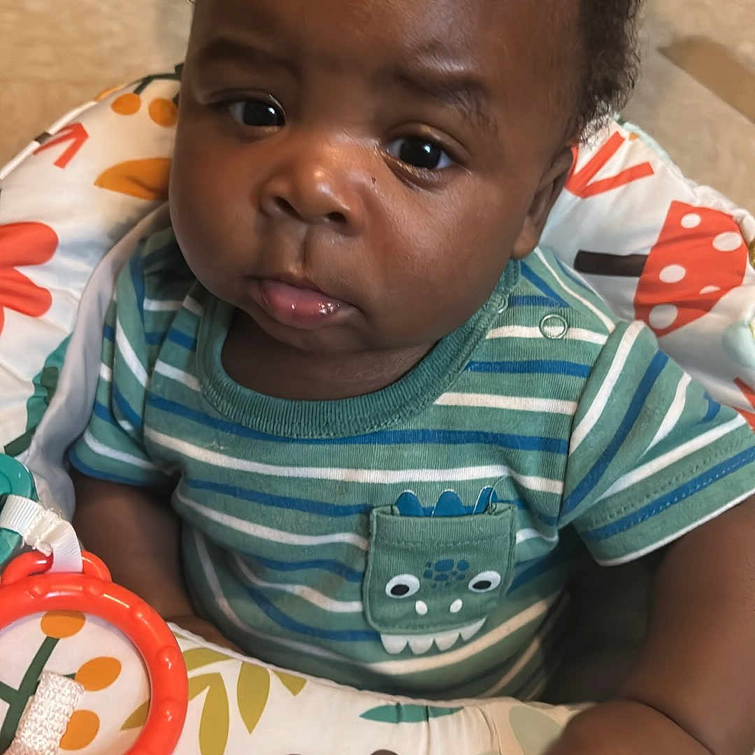 Cayden joined the competition — help win amazing prizes! baby, child, infant, striped_shirt, curly_hair, baby_seat, toy_ring, colorful, indoor, floor, bare_feet, cute, expression, portrait, person, young_child, playful, seated, hand, face