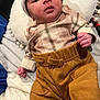 baby, infant, plaid_shirt, mustard_pants, hat, pillow, hand, face, clothing, newborn, lying_down, soft_texture, blanket, cute, indoors, portrait, child, expression, cozy, sleepwear