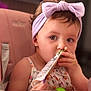 toddler, child, baby, headband, bow, high_chair, snack_pouch, feeding, indoor, cute, pink, clothing, person, face, hand, tableware, spoon, hair, earrings, expression