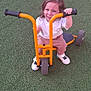 child, daylight, fun, green_turf, happy, orange, outdoor, person, pink_pants, play, playground, recreation, red_cone, smiling, sneakers, toddler, toy, tricycle, white_shirt, young_child
