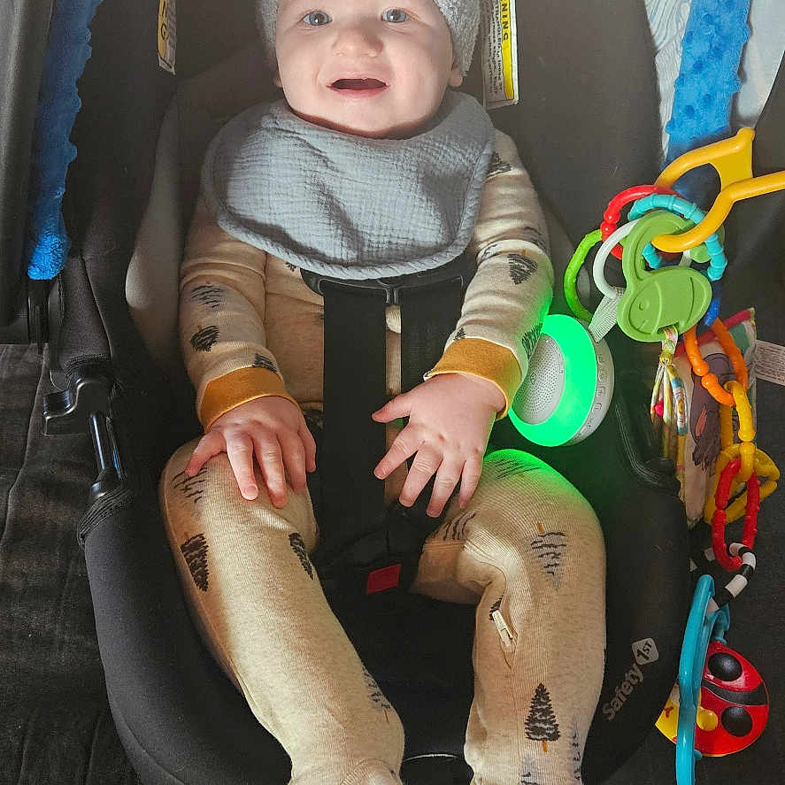 Cato is registered to the contest to win money with this photo: baby, blanket, car_seat, child, clothing, colorful_toys, cushion, face, footed_pajamas, hands, happy, hat, indoor, infant, pajamas, person, seatbelt, smiling, toy, wooden_floor