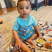 Eddie is registered to the contest to win money with this photo: toddler, child, baby, indoor, carpet, blanket, toy, bottle, clothing, person, face, curly_hair, floor, playpen, furniture, living_room, expression, sitting, casual, home