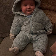 Theo joined the competition — help win amazing prizes! baby, child, onesie, blue_clothing, couch, pillow, soft_texture, indoor, cute, smiling, seated, hood, face, person, cozy, infant, warm_clothing, sock, plush, home