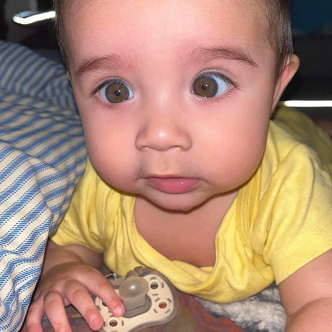 Elias joined the competition — help win amazing prizes! baby, child, face, eyes, pacifier, hand, yellow_shirt, blanket, indoor, portrait, curious, cute, young, infant, skin, hair, expression, lying_down, person, closeup