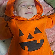 Juniper is registered to the contest to win money with this photo: baby, blanket, child, clothing, cozy, cute, fabric, festive, holiday, hood, indoors, infant, lying_down, orange_hoodie, person, portrait, pumpkin_face, sleepwear, smile, soft