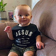 Huxon is registered to the contest to win money with this photo: baby, child, smiling, sitting, couch, clothing, black_shirt, camouflage_shorts, indoor, plant, decor, face, happy, person, foot, hand, home, furniture, portrait, toddler