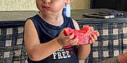 Huxon is registered to the contest to win money with this photo: arm, background, checkered, child, clothing, expression, face, food, hair, indoor, person, shorts, sitting, smartphone, stool, table, tank_top, tattoo, toddler, watermelon