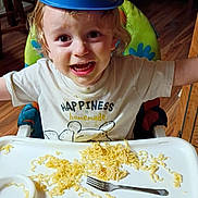 Huxon is registered to the contest to win money with this photo: child, toddler, highchair, noodles, fork, bowl, hat, tray, tshirt, smile, happy, messy, food, face, hair, indoor, wooden_floor, chair, dining_room, person