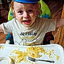 child, toddler, highchair, noodles, fork, bowl, hat, tray, tshirt, smile, happy, messy, food, face, hair, indoor, wooden_floor, chair, dining_room, person