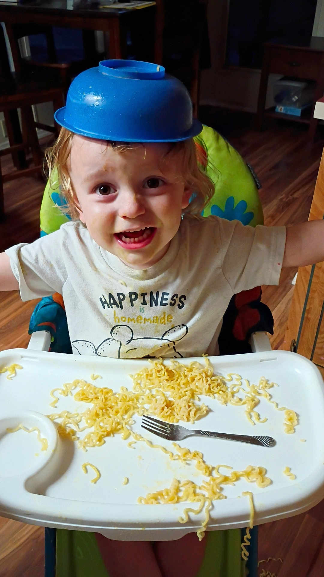 Huxon is registered to the contest to win money with this photo: child, toddler, highchair, noodles, fork, bowl, hat, tray, tshirt, smile, happy, messy, food, face, hair, indoor, wooden_floor, chair, dining_room, person