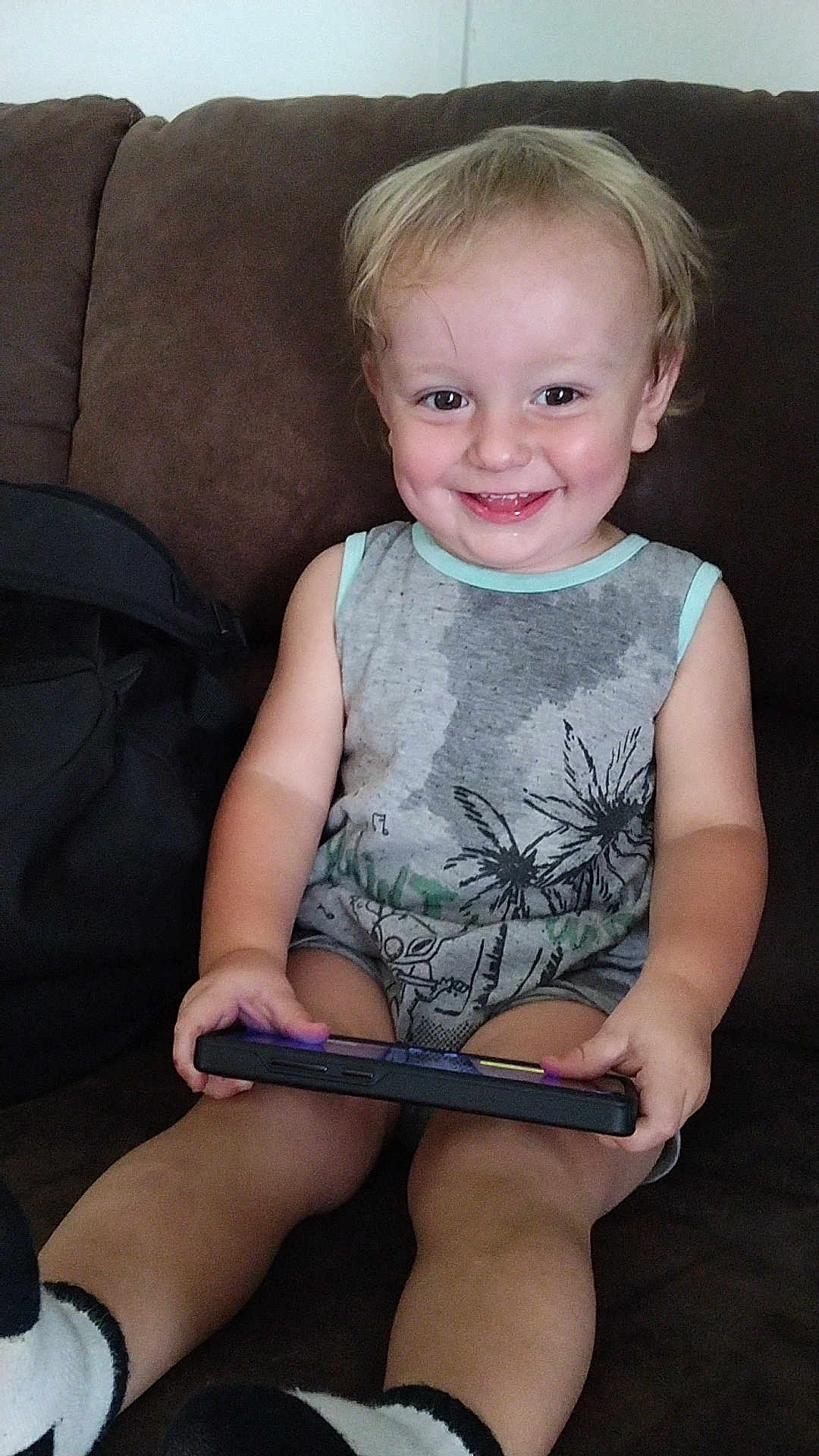 Huxon is registered to the contest to win money with this photo: toddler, child, smiling, sitting, couch, brown_couch, handheld_device, gaming, socks, casual_clothing, indoor, happy, light_hair, eyes, person, young_child, furniture, relaxed, playful, portrait