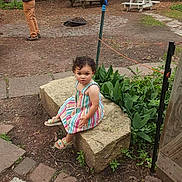 Lydia Altamirano is registered to the contest to win money with this photo: toddler, child, outdoor, garden, stone_block, striped_dress, curly_hair, greenery, picnic_table, person, pathway, plant, sandals, serious_expression, daylight, nature, casual, playground, young_child, sitting