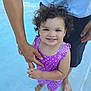 child, toddler, curly_hair, blue_eyes, purple_outfit, polka_dot, smiling, barefoot, water, pool, adult_hand, support, summer, outdoor, cute, happy, skin, person, family, play