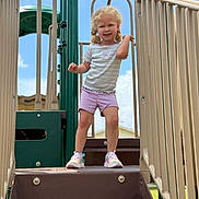 Nyomi joined the competition — help win amazing prizes! child, playground, smiling, curly_hair, shorts, striped_shirt, flower_emblem, sneakers, outdoor, blue_sky, clouds, play_structure, railing, steps, happy, person, daylight, summer, park, young_child