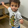 toddler, child, curly_hair, indoor, table, tshirt, teenage_mutant_ninja_turtles, shorts, floor, wooden_table, brown_eyes, person, young_child, casual_clothing, standing, looking_up, lip_bite, cute, curious, light_background